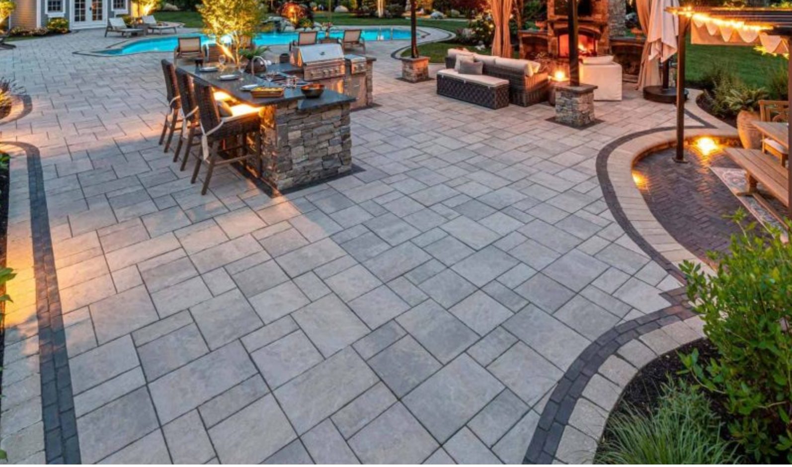 Unilock paver patio at night with outdoor kitchen, pool and fireplace lounge